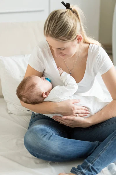 Beautiful young woman sitting on bed na drocking her sleeping baby boy — Stock Photo, Image