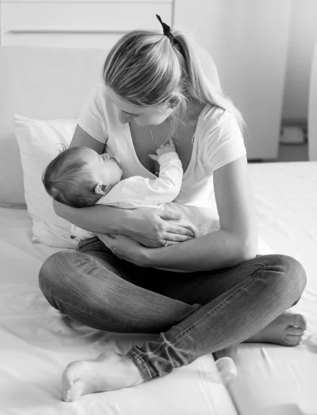 Schwarz-Weiß-Foto einer jungen Frau, die auf dem Bett sitzt und ihr schlafendes Baby hält — Stockfoto