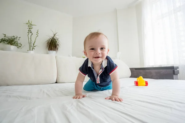 Широкоугольное фото улыбающегося мальчика ползающего по кровати и смотрящего в камеру — стоковое фото