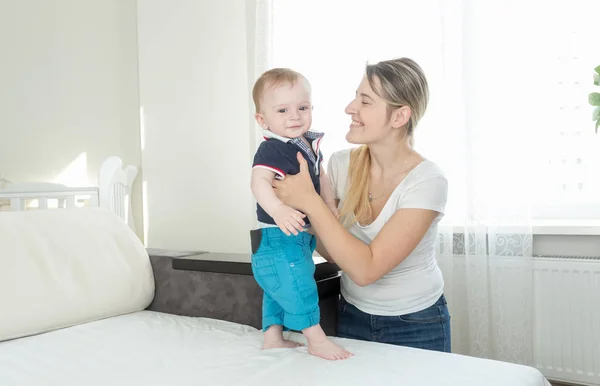 Retrato de feliz madre sonriente sosteniendo a su hijo bebé de pie en el sofá —  Fotos de Stock