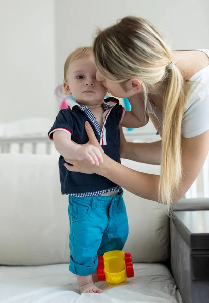 Portret młodej kobiety, całując jej synka, stojąc na kanapie w sypialni — Zdjęcie stockowe