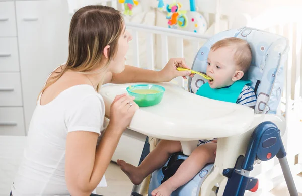 Retrato de menino de 10 meses sentado em cadeira alta e comendo mingau de cereal de colher — Fotografia de Stock