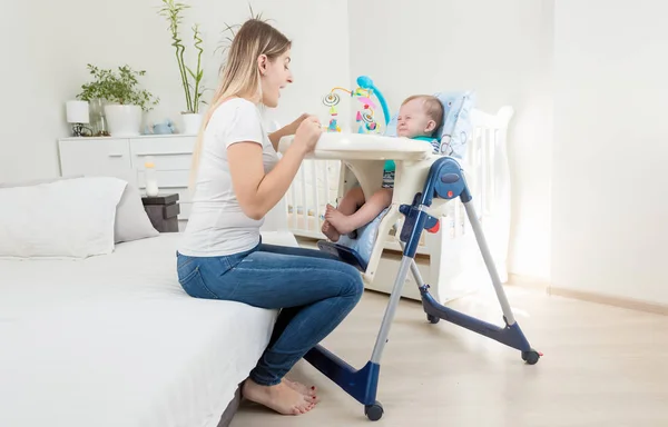 Jovem mãe tentando alimentar seu bebê chorando sentado em cadeira alta — Fotografia de Stock