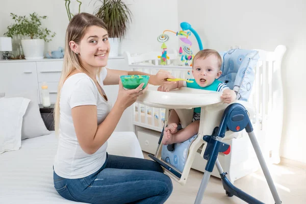 Retrato de una joven madre sonriente alimentando a su hijo con el pecado en la trona y mirando a la cámara — Foto de Stock