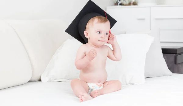 Lindo niño en pañales y sombrero de graduación sentado en el sofá en casa — Foto de Stock