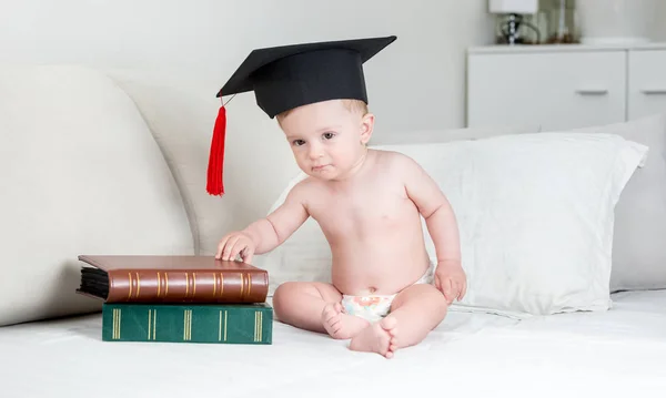 Söt baby boy i blöjor och examen mössa sitter på sängen med böcker — Stockfoto