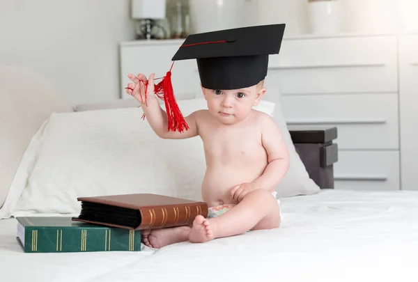 Retrato de niño inteligente en gorra de mortero sentado con libros en el dormitorio — Foto de Stock