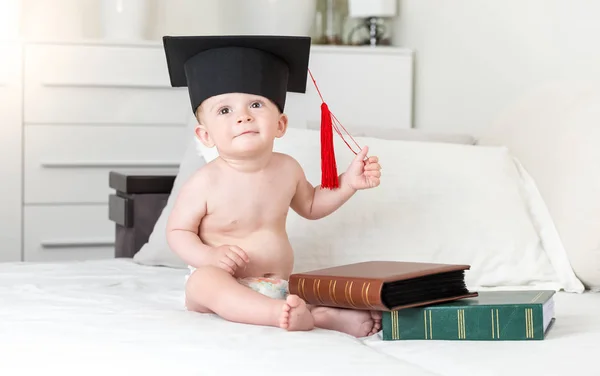 Entzückend lächelnder Kleinkind-Junge mit Mörtelmütze und Büchern auf dem Bett — Stockfoto