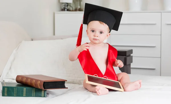 Cute toddler boy in graduation hat and red ribbon holding books and blackboad — Stock Photo, Image