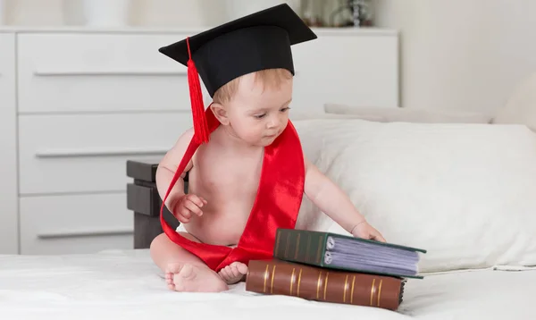 Bonito bebê menino no preto graduação chapéu com tissel olhando para Grande idade livros — Fotografia de Stock