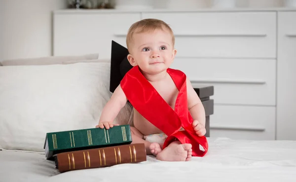 Portrait de bébé garçon intelligent dans le ruban rouge de graduation posant avec des livres sur le lit — Photo