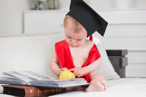 Retrato de lindo bebé niño en gorra de graduación sosteniendo manzana y libro de lectura. Concepto de educación infantil temprana — Foto de Stock