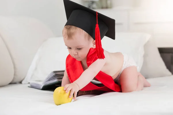 Retrato de bebé niño en pañales y sombrero de graduación con tissel rojo arrastrándose en la cama hacia la manzana grande. Concepto de niños inteligentes — Foto de Stock