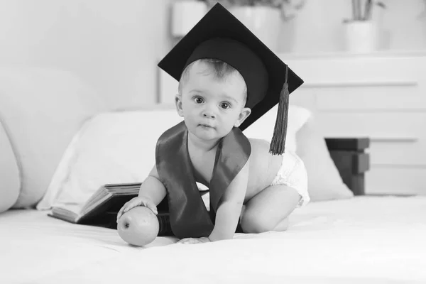 Imagen en blanco y negro del niño genio en sombrero de graduación sentado en la cama con manzana y mirando a la cámara — Foto de Stock