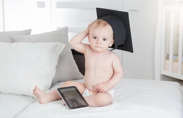 Rolig bild av bedårande pojke i blöjor och examen hatt sitter på sängen med digital tablett — Stockfoto