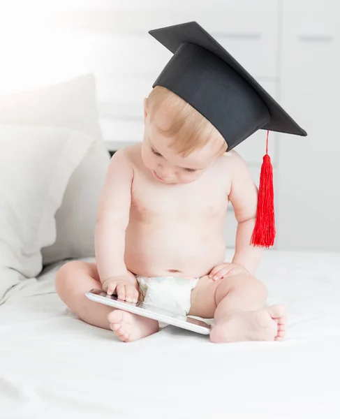 Söt baby boy i examen cap sitter på sängen och använder digital tablett dator — Stockfoto
