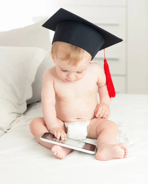 Porträtt av lilla pojke i blöjor bär examen cap med på sängen och använda digitala tablett — Stockfoto
