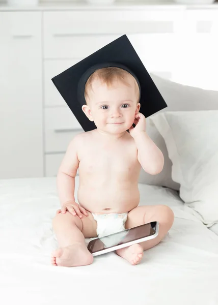 아기 기저귀 입고 대학 졸업에에서 미소의 모자 침대에 앉아서 태블릿 컴퓨터를 사용 하 여 — 스톡 사진