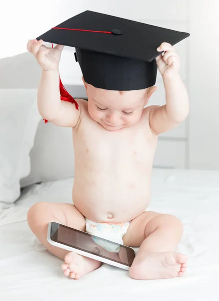 Porträtt av pojke i blöjor och examen mössa sitter på sängen och surfar internet på digitala tablett. Begreppet smarta barnen och tidig utbildning med användning av modern teknik och datorer — Stockfoto