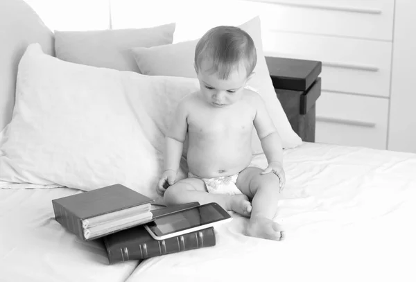Immagine in bianco e nero di adorabile bambino in pannolini seduto sul letto con grande atck di libri e tablet digitale. Concetto di bambini intelligenti e istruzione precoce con l'uso di tecnologie moderne e computer — Foto Stock