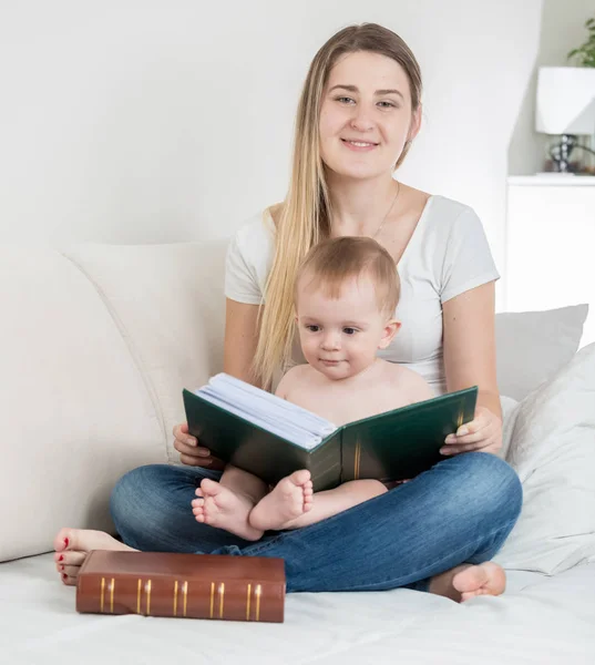 Bela jovem mãe com bebê menino sentado na cama e segurando grande livro velho — Fotografia de Stock