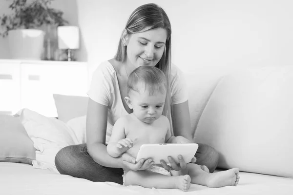 Черно-белый образ молодой мышиной матери, сидящей со своим малышом на кровати и с помощью цифрового планшета — стоковое фото