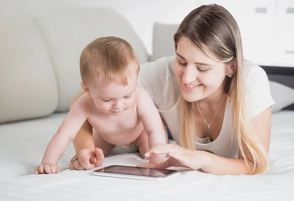 Тонированный портрет улыбающейся матери с ребенком, лежащим на кровати и просматривающим интернет на цифровом планшете — стоковое фото