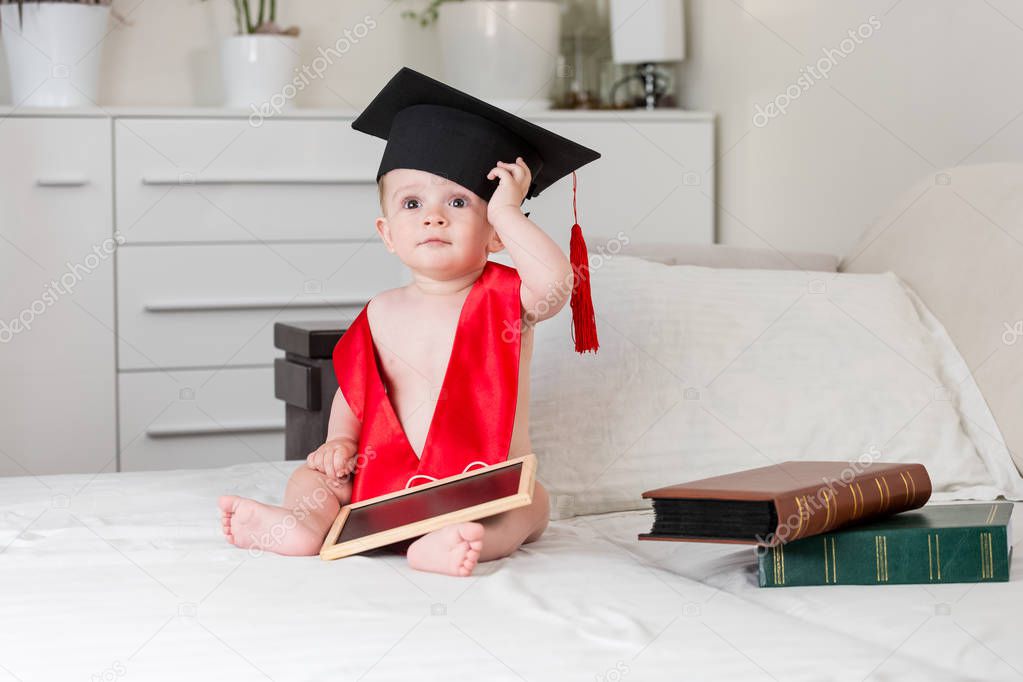 Cute baby boy in graduation hat and red collar sitting on bed with books and chalkboard