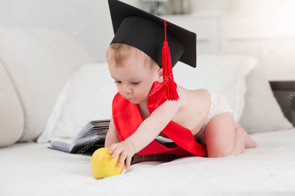 Portrait of baby boy in diapers and graduation hat with red tissel crawling on bed towards big apple. Concept of smart kids
