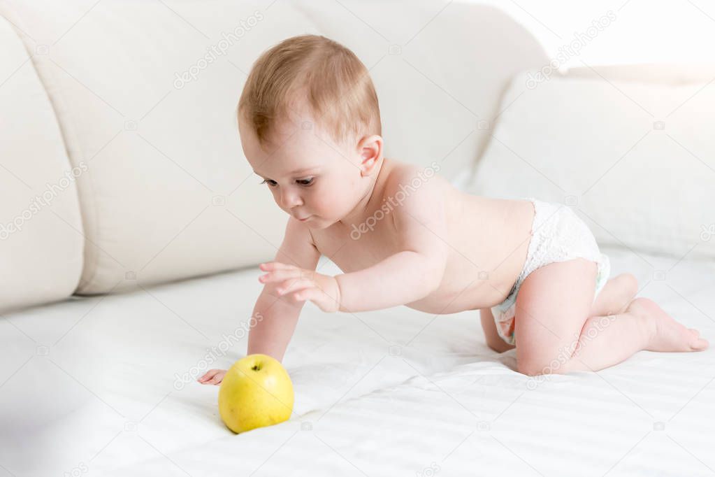Cute baby boy in diapers crawling on bed and reaching for big yellow apple