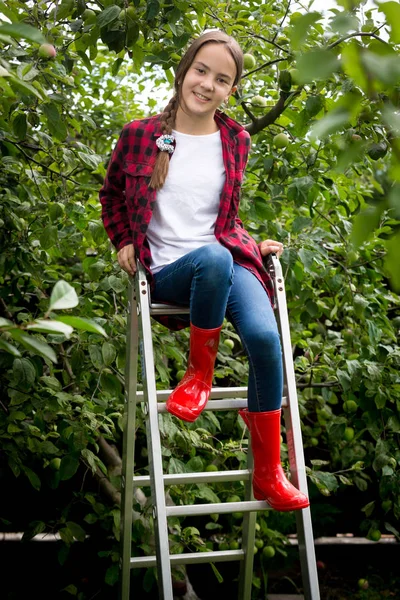 Beautiful smiling girl in red rubber boots sitting on stepladder at garden — Stock Photo, Image