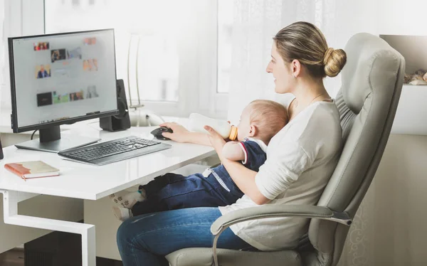 Young woman working at home with computer and feeding her baby with milk — Stock Photo, Image