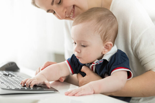 Cute baby boy sitting on mothers lap in office and typing on computer keyboard