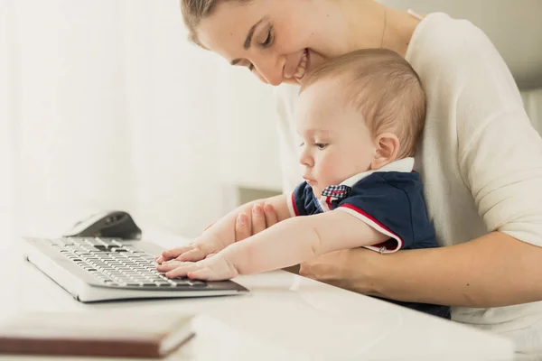 Тонированный портрет улыбающейся молодой матери, обучающей своего маленького сына с помощью компьютера в домашнем офисе — стоковое фото