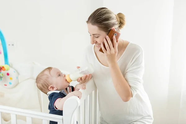 Cute baby boy eating milk from bottle and looking at mother talking by phone — Stock Photo, Image