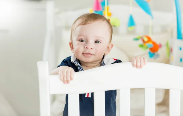 Portrait of cute baby boy standing in white wooden crib and looking in camera — Stock Photo, Image