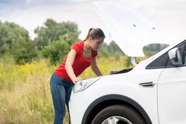 Portrait of upset young woman opened hood looking at engine of broken car — Stock Photo, Image