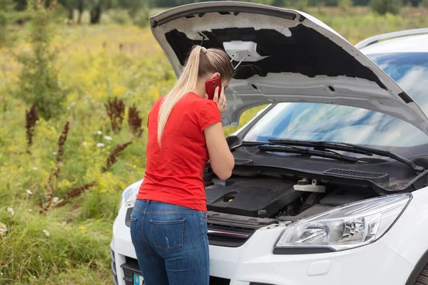 Rear view image of young woman looking under the hood of broken car and calling service — Stock Photo, Image