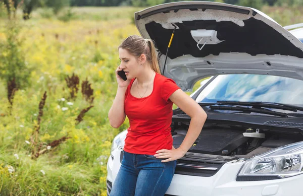 Portrait of young upset woman leaning on broken car in field and calling for help — Stock Photo, Image