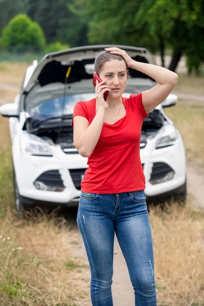 Portrait of stressed young woman standing at her broken car and calling car service — Stock Photo, Image