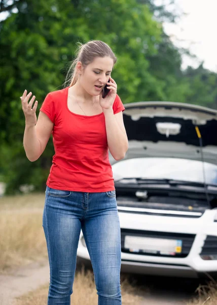 Portret van boze jonge vrouw praten via de telefoon en aanroepen van bijstand aan haar gebroken auto in veld — Stockfoto