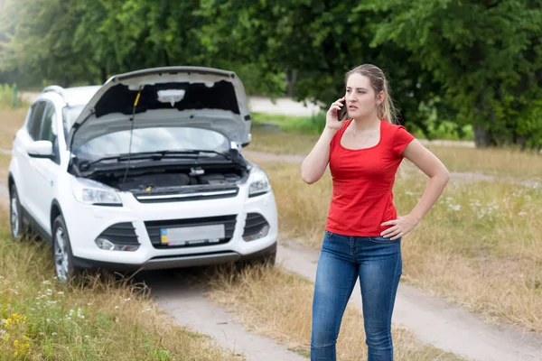 Jeune femme en colère debout à sa voiture cassée dans le champ et parlant par téléphone avec le mécanicien — Photo