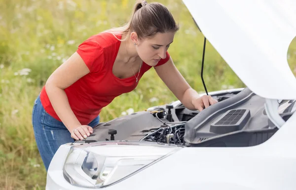 Portrait of upset young woman looking under the hood of broken car and trying to fix engine — Stock Photo, Image