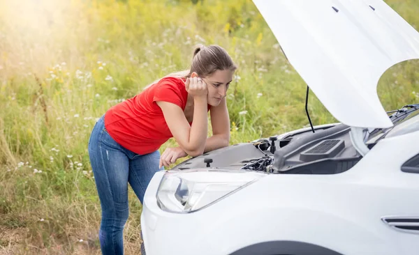 Upset young woman leaning on broken car with open hood in field — Stock Photo, Image