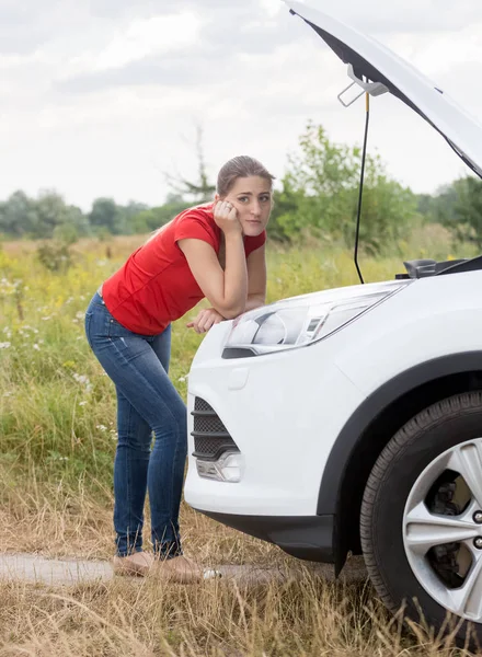 Upset young woman leaning on broken car in field looking in camera — Stock Photo, Image