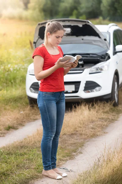 Young woman standing at her broken car in field and reading instructions book — Stock Photo, Image
