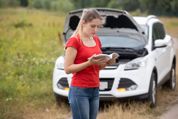 Portrait of stressed young woman reading manual instructions for her broken car — Stock Photo, Image