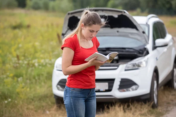 Portrait of stressed young woman with broken car reading manual — Stock Photo, Image
