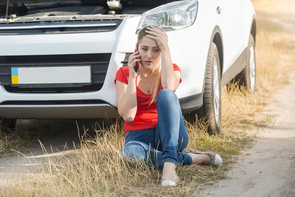 Retrato de una joven trastornada sentada en el suelo junto a su coche roto y hablando con el servicio por teléfono móvil —  Fotos de Stock