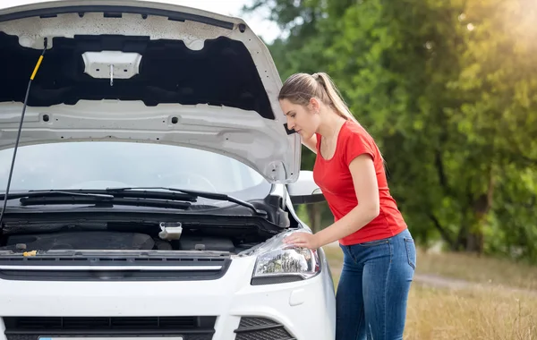 Расстроенная молодая женщина смотрит на перегретый двигатель своей сломанной машины — стоковое фото